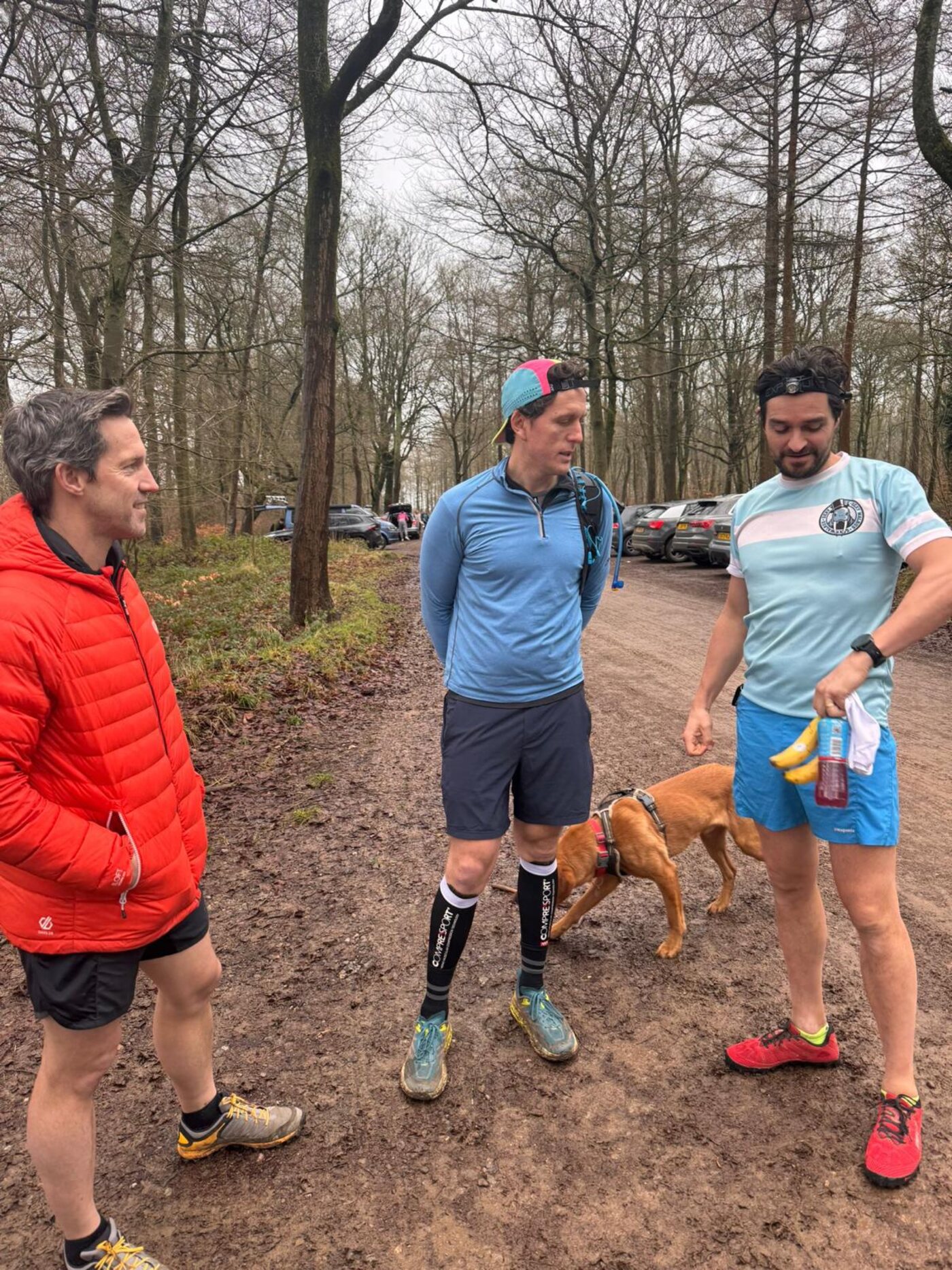 Three club members talking in a muddy forest car park before a winter run, one in club kit, a dog beside them