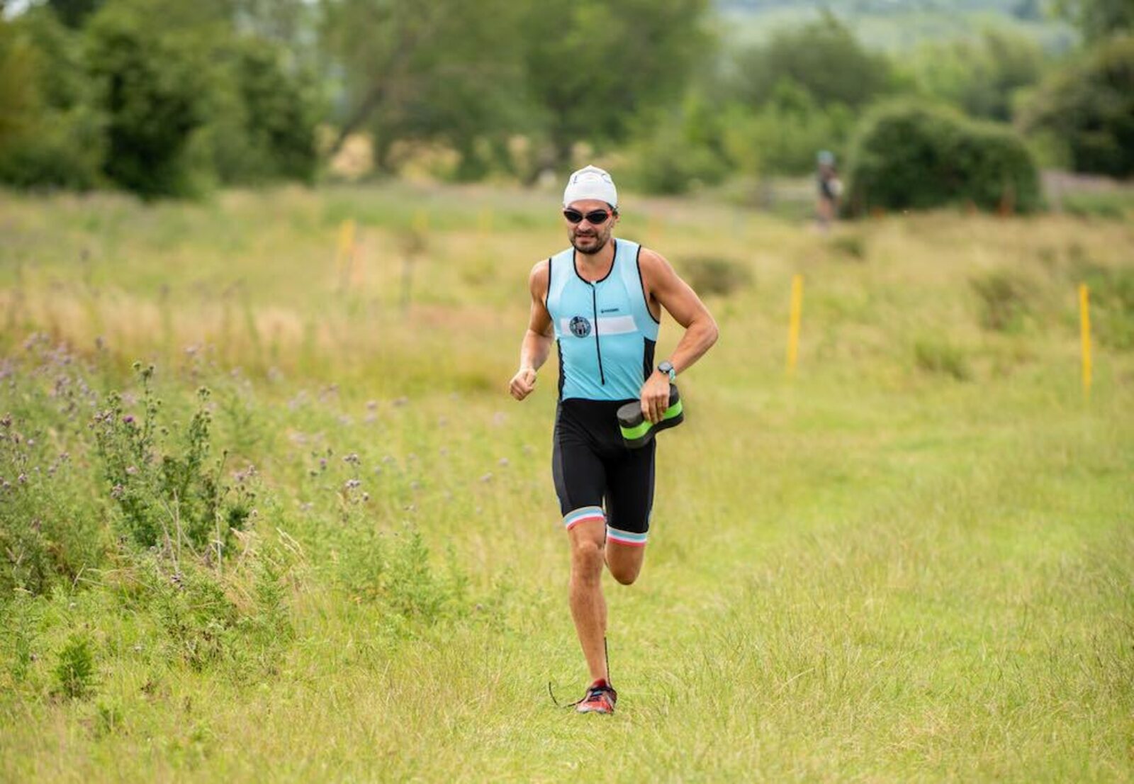 A club member running on grass in club kit, carrying his shoes, with course markers behind him