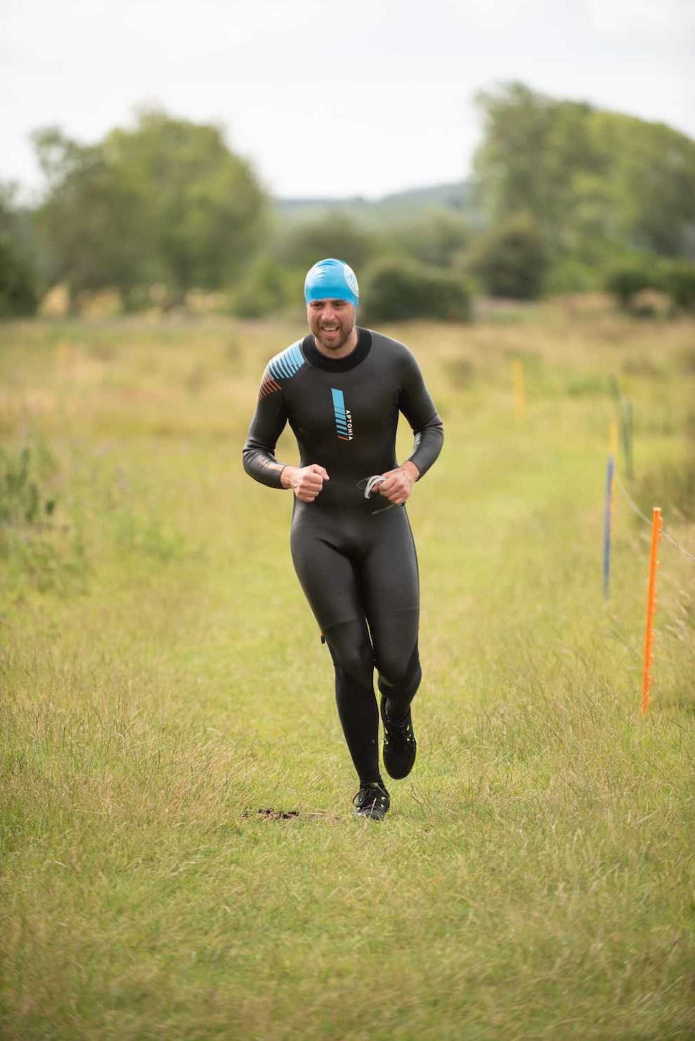 A member running on grass in a wetsuit with a swim cap on, goggles in hand, course markers behind