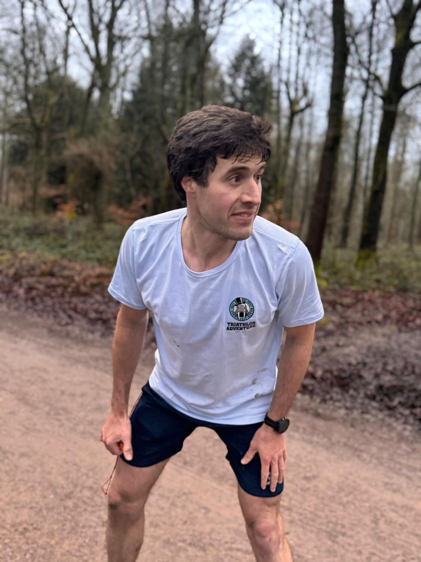 A member in a mud-spattered club t-shirt leaning over with his hands on his knees in a winter wood